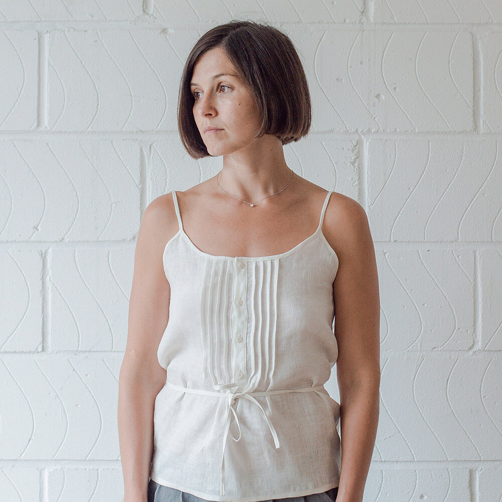 Woman wearing a white linen camisole with pintuck detailing, buttons down the front, and waist tie.