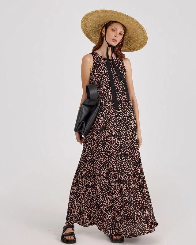 Woman wearing a sleeveless leopard print maxi dress with black tie detail, wide-brim straw hat, and sandals.