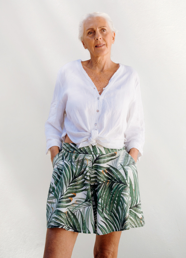 Woman wearing a white button-up blouse and green tropical print shorts, standing against a white wall.