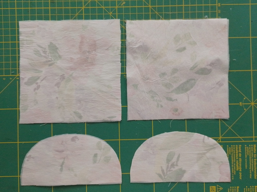 Two square fabric pieces and two curved flap pieces with interfacing applied, laid out on a cutting mat.
