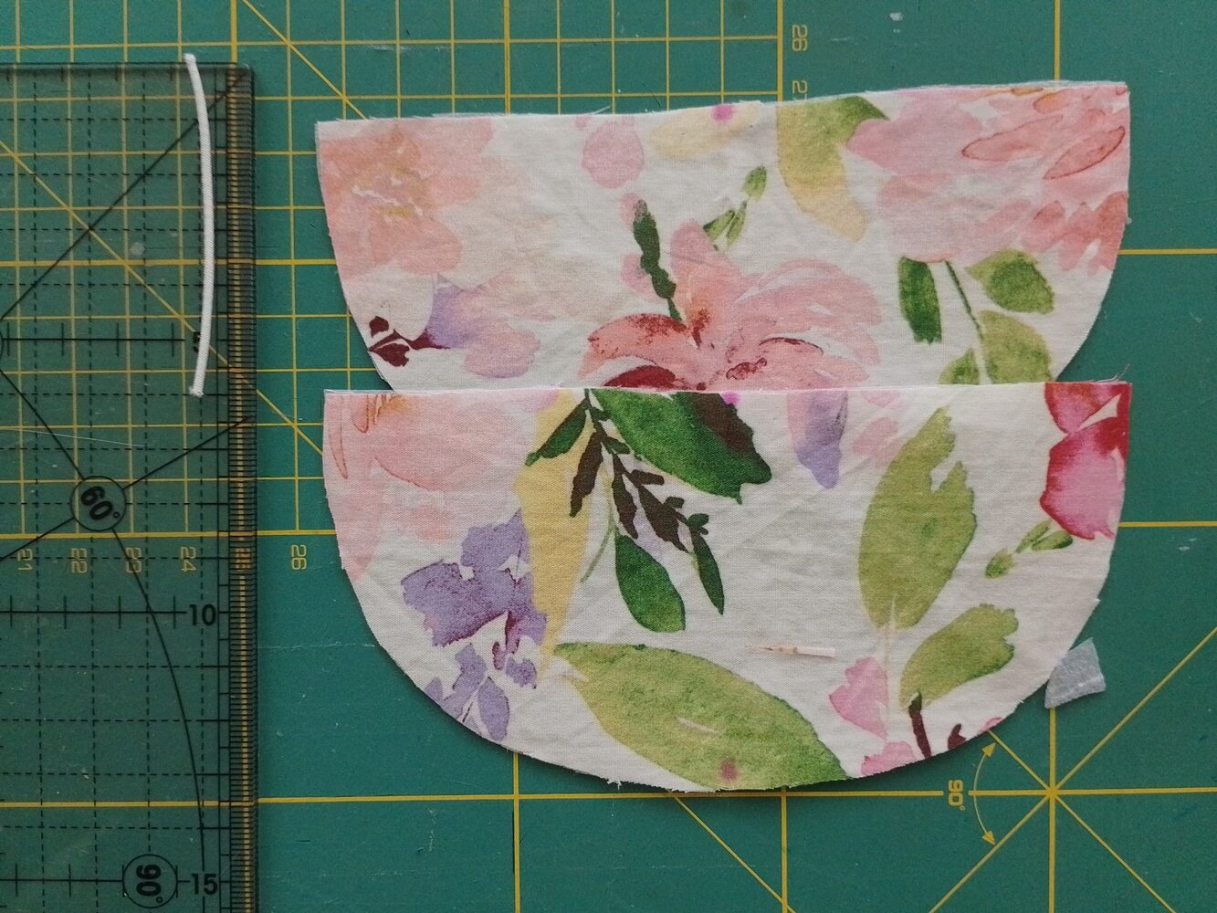 Two curved flap fabric pieces with floral print and a 6 cm piece of cord elastic placed next to a ruler on a green cutting mat.