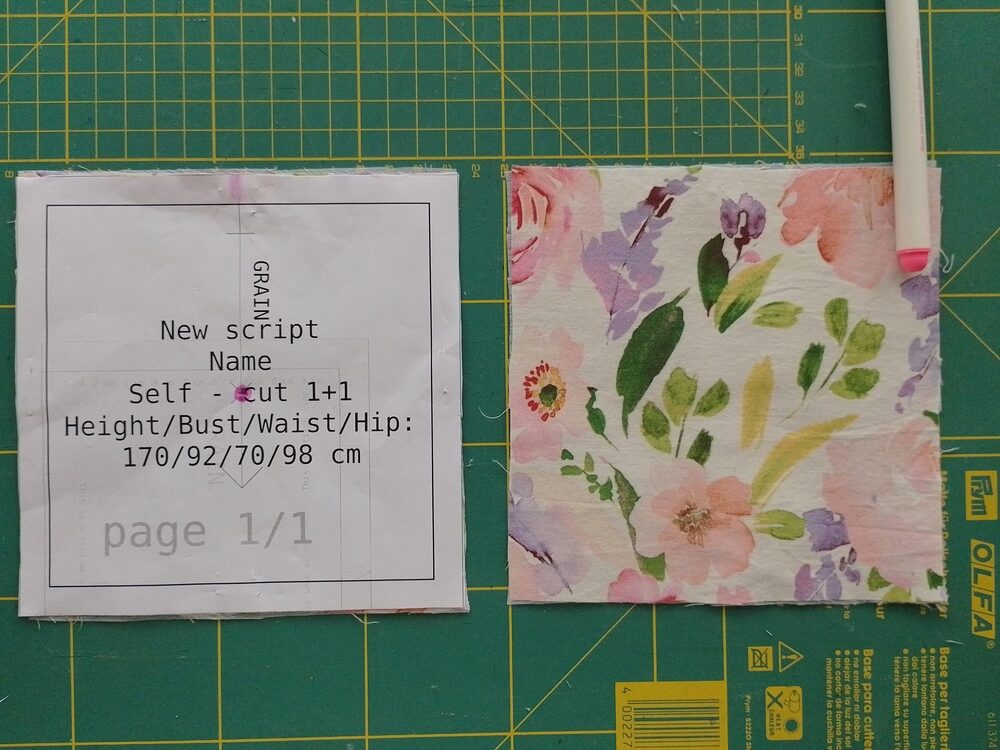 Fabric square next to paper pattern with a center X marked for button placement. These and the end of a marker pen are on a green cutting mat.