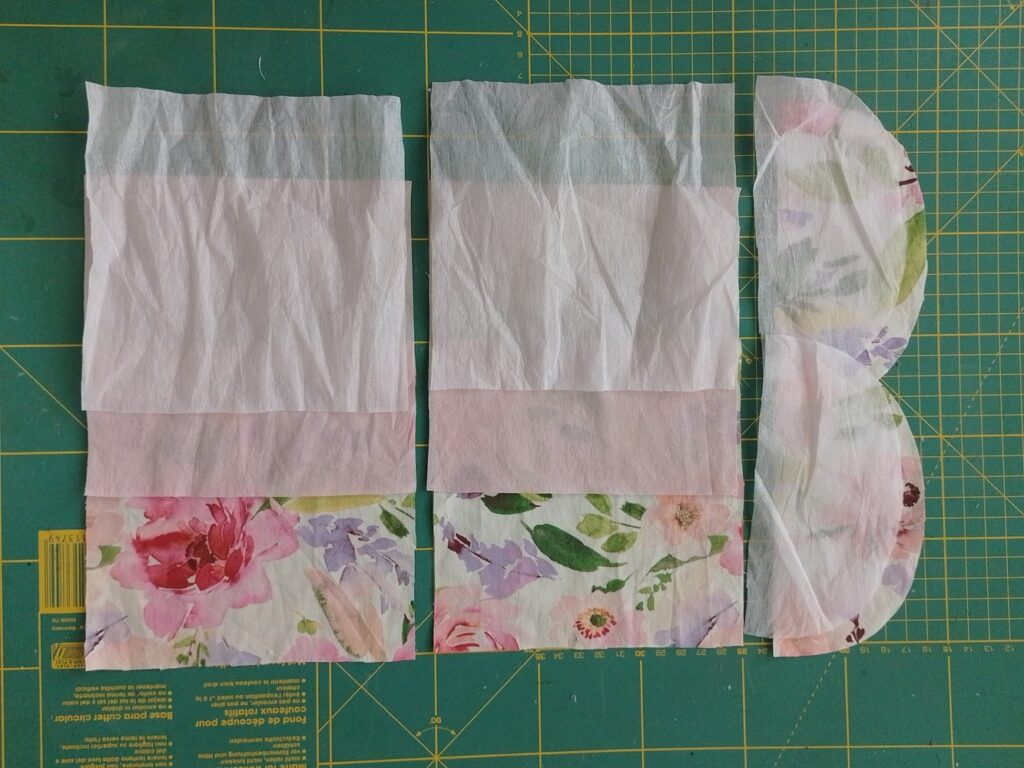 Cut fabric pieces for a sanitary pad pouch, including two rectangles and two curved flap pieces, laid out on a cutting mat.