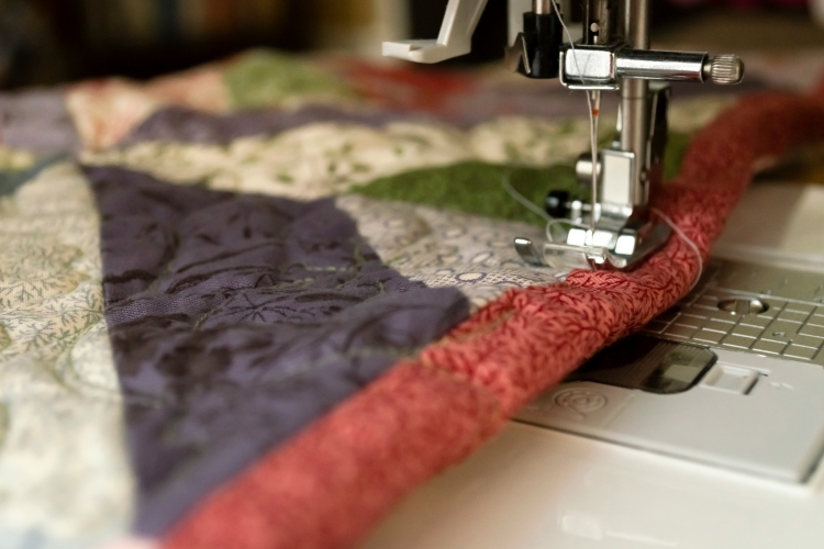 A close-up view of a sewing machine's needle and presser foot stitching binding onto a quilt.