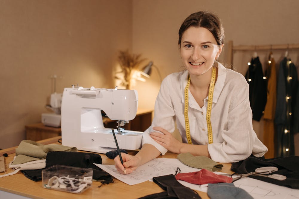 Query successful
A smiling woman with a measuring tape around her neck sits at a table with a sewing machine, sketching designs on paper. Various fabric swatches and sewing tools are also on the table.