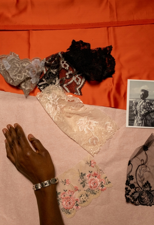 An overhead shot of a designer's workspace shows colorful fabric, various lace samples, and an inspirational vintage black-and-white photograph. A person's hand, wearing a silver bracelet, rests beside the materials.