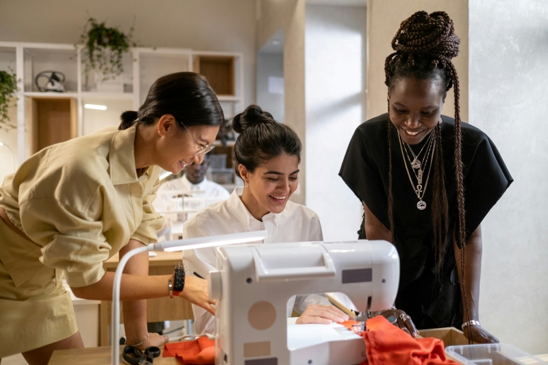 Three women are gathered around a sewing machine, smiling.
