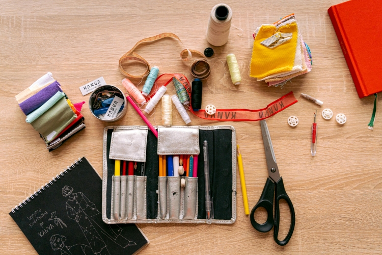 A flat lay of sewing and fashion design supplies on a light wooden surface. In the center is an open, silver roll-up pencil case filled with colored pencils. Surrounding it are a pair of black-handled scissors, spools of thread in various colors, a measuring tape, stacks of colorful fabric swatches, a seam ripper, buttons, and a sketchbook with fashion illustrations on the cover.