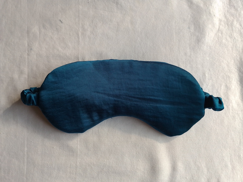 The front of a teal satin eye mask lying flat on a cream-colored fabric background.