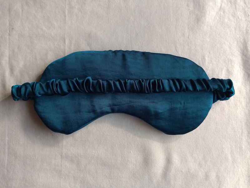 The back view of a teal eye mask, showing the ruched fabric strap stretching horizontally across the center of the mask.