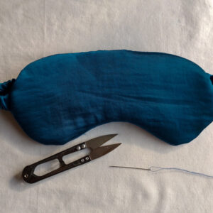 The finished teal satin eye mask on a cream background, with a pair of black thread snips and a threaded needle below it.