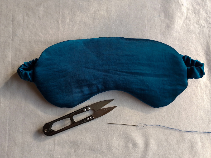 The finished teal satin eye mask on a cream background, with a pair of black thread snips and a threaded needle below it.