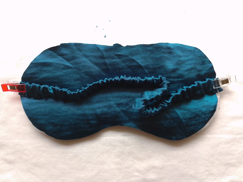 The teal, ruched fabric strap is positioned horizontally across the front of the eye mask. A red sewing clip on the left and a clear sewing clip on the right secure the ends of the strap to the mask.