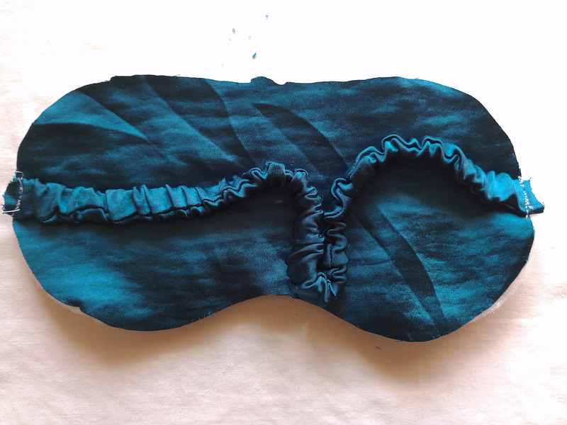 The teal fabric strap is now sewn onto the sides of the eye mask, with white stitches visible at both attachment points.
