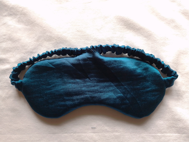 A teal satin eye mask, now right-side out, lying on a cream-colored surface. The fabric is slightly wrinkled and the ruched strap is attached. There is an open gap at the top of the sleep mask
