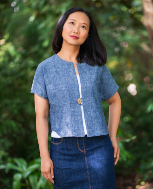 A woman models a short-sleeved, open-front topper made of blue fabric, worn over a white shirt and denim skirt.