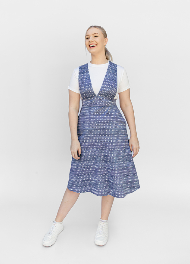 A smiling woman models a blue patterned V-neck pinafore dress layered over a white t-shirt.