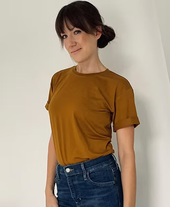 A woman with brown hair in a low bun with bangs smiles while modeling a mustard-yellow t-shirt. The short sleeves are cuffed, and the shirt is tucked into blue jeans. She is standing against an off-white wall.
