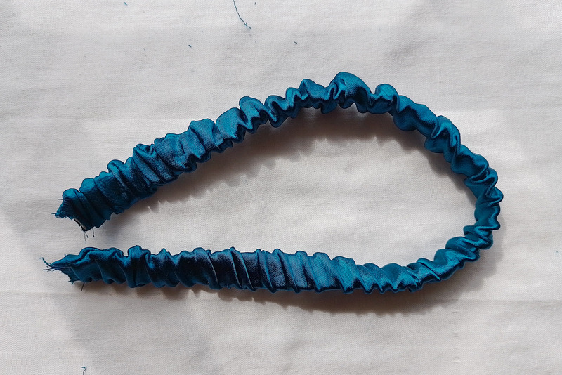 The teal fabric strap that is gathered and ruched, lying in a loop on a white surface.