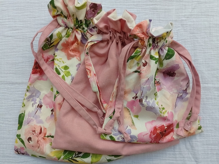 A set of three matching drawstring bags in various sizes are nestled together on a white background. The bags are sewn from a coordinating floral print and solid pink fabric, each with a ruffled top.