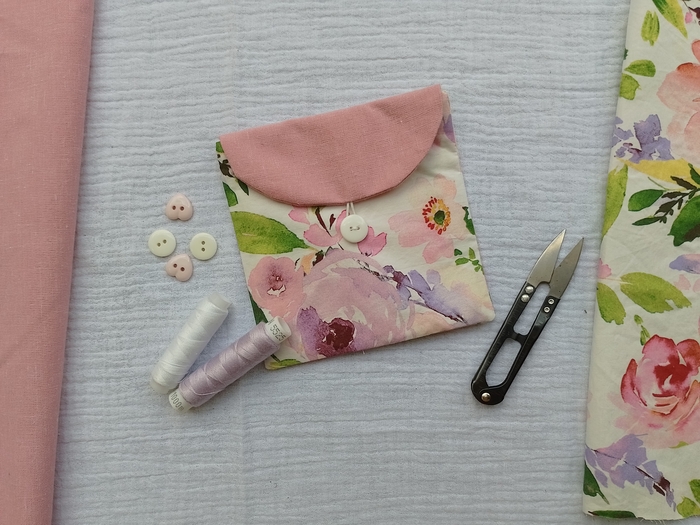 A small, handmade floral pouch with a pink flap and white button is displayed on a white textured fabric. Surrounding the pouch are sewing supplies, including small black scissors, spools of white and lilac thread, and several heart-shaped buttons.