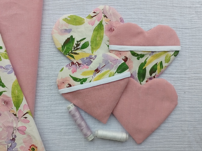 Four handmade, heart-shaped coasters are displayed on a white background. They show different quilted designs: one is solid pink, one is floral, and two are a patchwork of both pink and floral fabric. Spools of thread and fabric swatches are arranged next to them.