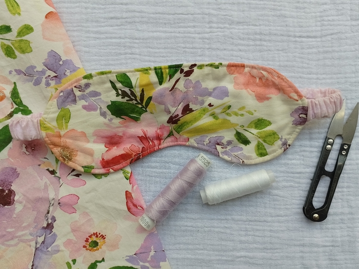 A handmade floral sleep mask with a ruched, pink fabric-covered strap lies on a white textured background. Next to it are thread snips and spools of white and lilac thread.