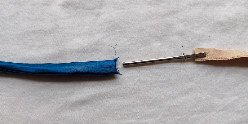 A blue fabric tube on the left, and on the right, a piece of beige elastic attached to a long metal bodkin tool.