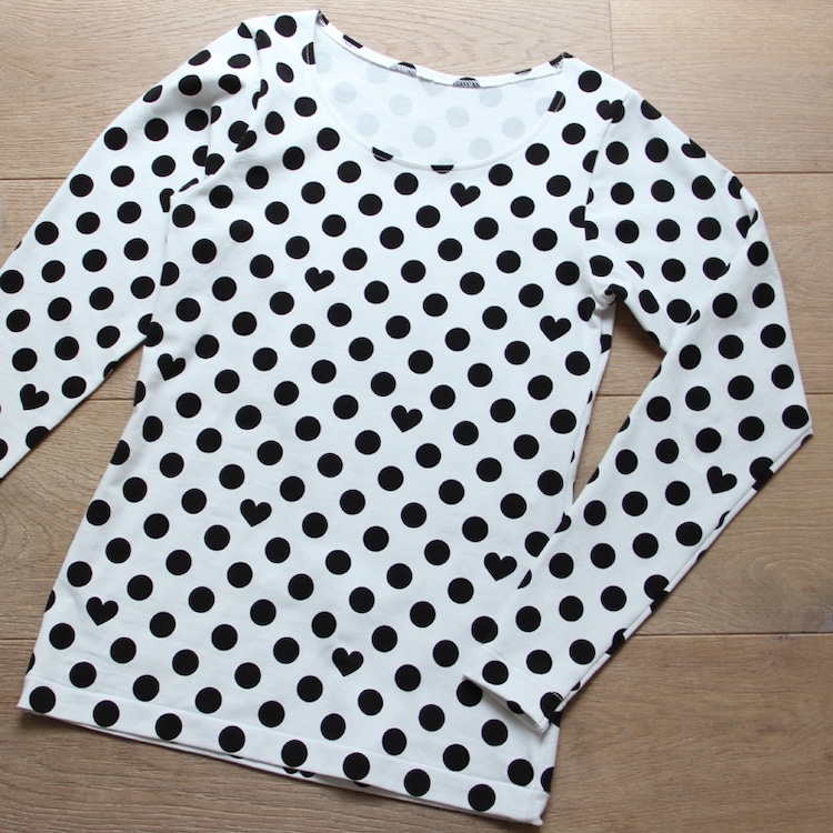 A white, long-sleeved t-shirt with a pattern of large black polka dots and small black hearts is laid flat on a light brown wooden surface.