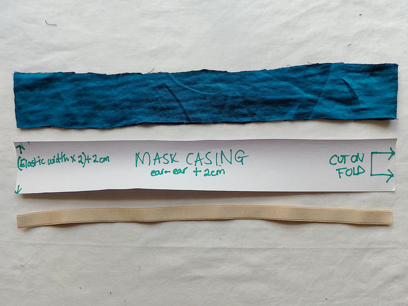 Three items for a mask strap laid out on a white surface: a rectangular piece of teal fabric, a paper pattern piece labeled "Mask Casing," and a piece of beige elastic.
