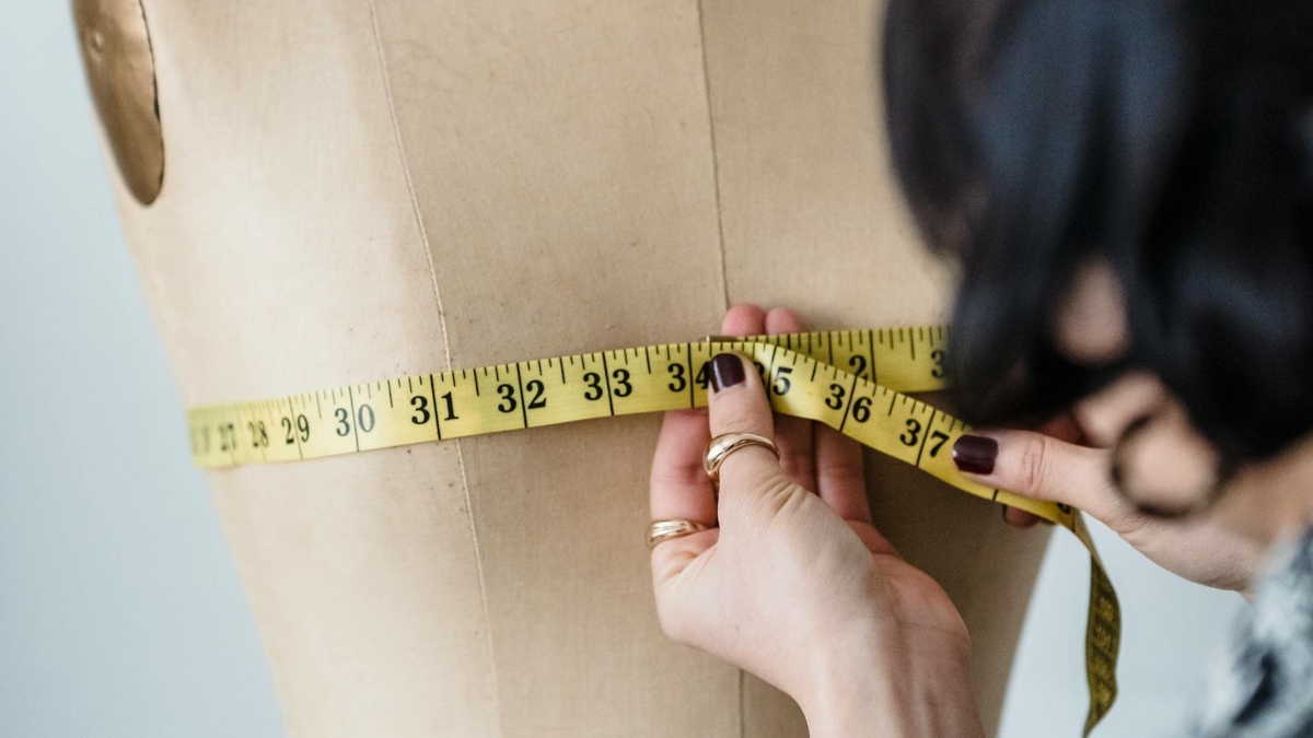 A woman's hands measuring the bust of a dress form with a yellow tape measure.
