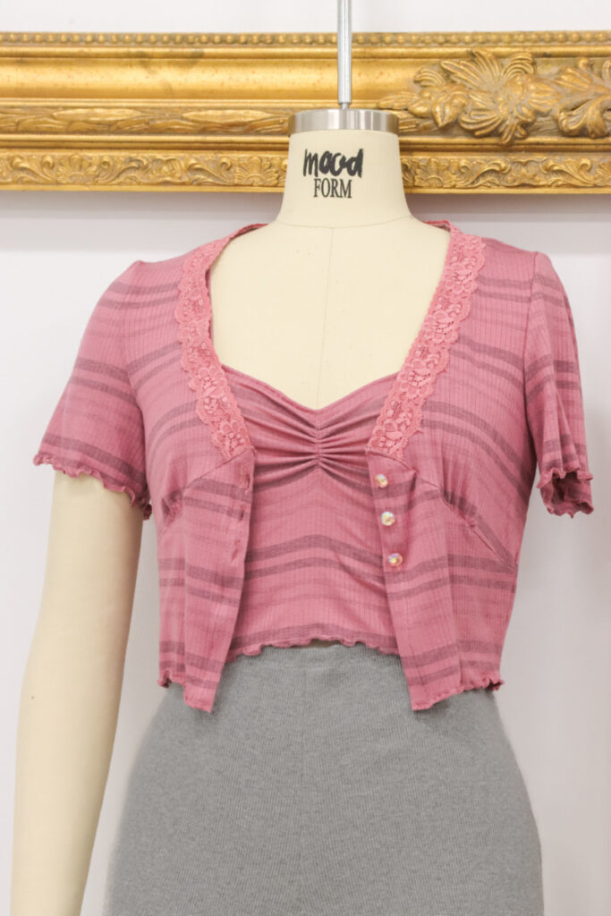 A cropped, short-sleeve pink cardigan with a lace-trimmed V-neck and button front, shown on a mannequin.