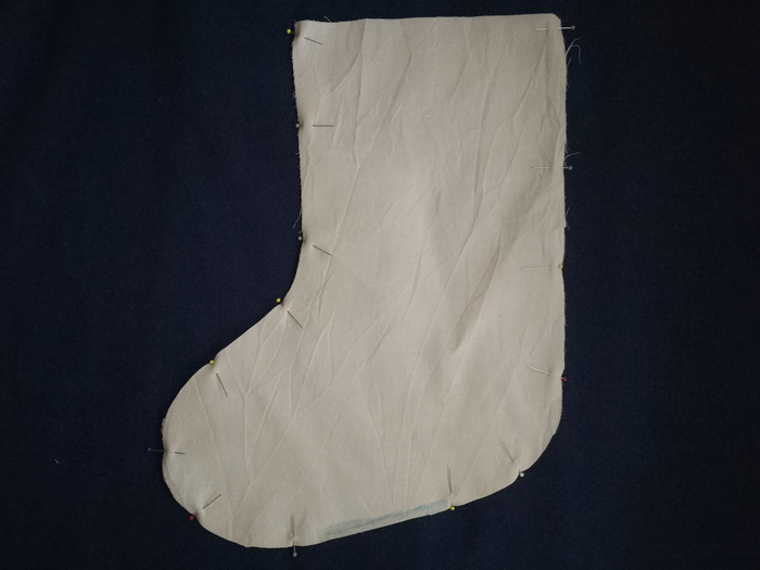 Two stocking-shaped pieces of off-white calico are pinned together around the edges against a dark blue background. A blue line is marked along the bottom curve.