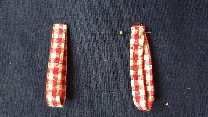 Two folded loops of red and white gingham fabric lying side-by-side on a dark blue background. The loop on the right has a green-headed pin through its top layers.