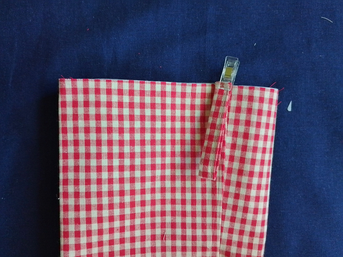A close-up showing a folded loop of red and white gingham fabric secured to the top edge of a gingham stocking with a clear sewing clip.