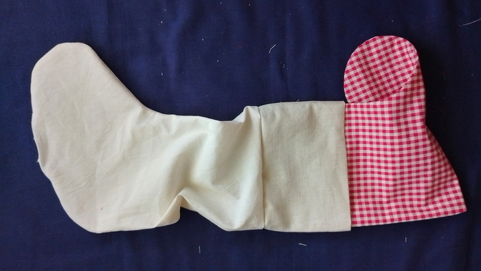 A red and white ginham stocking, off white calico lining and off white linen cuff turned right-side out and laid flat, connected at the top seam, on a dark blue background.