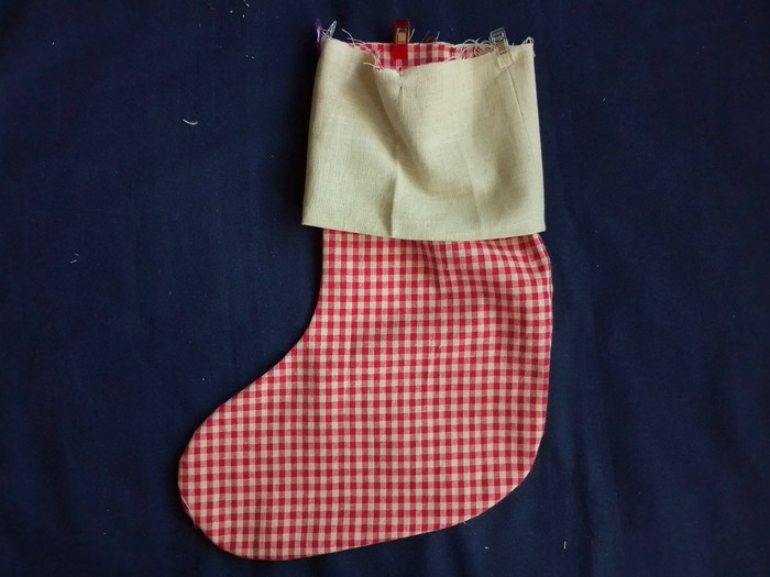 An off-white linen cuff secured with sewing clips around the top opening of a red and white gingham stocking.
