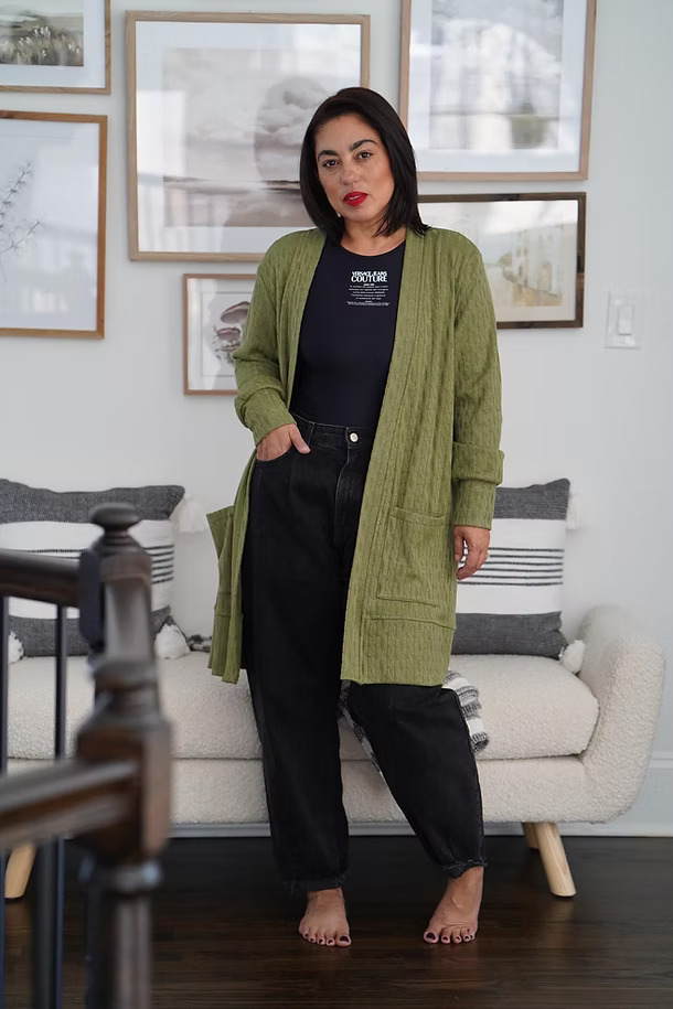 A woman wearing a long, olive green open-front cardigan made from a textured ribbed knit, paired with black jeans.