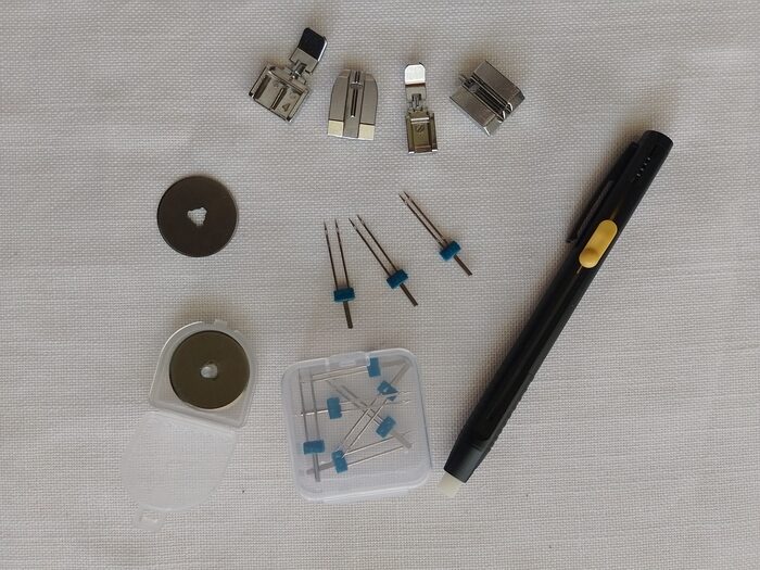 A flat lay of assorted sewing machine tools including four presser feet, twin needles, a case of sewing needles, rotary cutter blades, and a black sewing chalk dispenser pen.