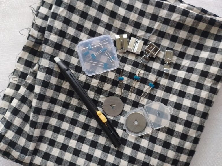 A variety of sewing machine presser feet, twin needles, rotary cutter blades, and a marking pen arranged in a cluster on top of checkered gingham fabric.