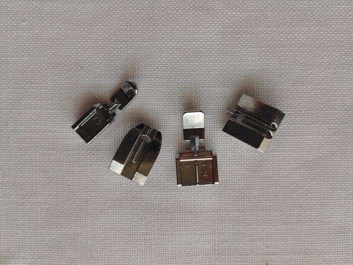 Four metal zipper presser feet for a sewing machine arranged on a light background.