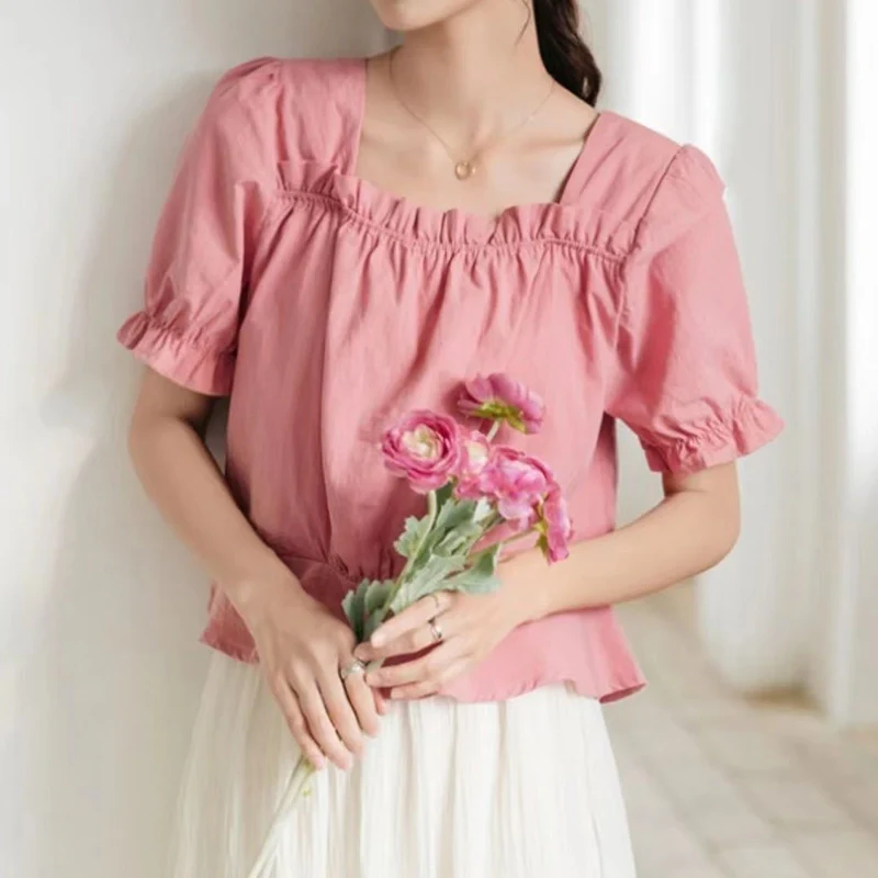 A person holding pink flowers, wearing a pink blouse with a square, ruffled elastic neckline and short puff sleeves.