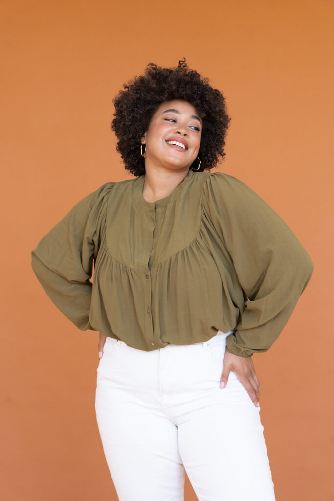 A smiling woman wearing a loose, olive green blouse with a gathered yoke and long sleeves, tucked into white pants.