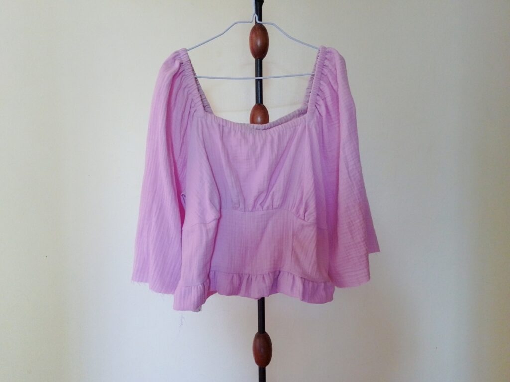 A lilac long-sleeved gathered top (Briarwood Top mock up) with a square neckline and ruffled hem, hanging on a hanger on a stand.