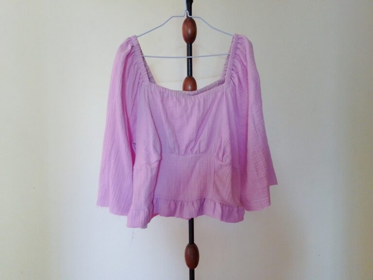 A lilac long-sleeved gathered top (Briarwood Top mock up) with a square neckline and ruffled hem, hanging on a hanger on a stand.
