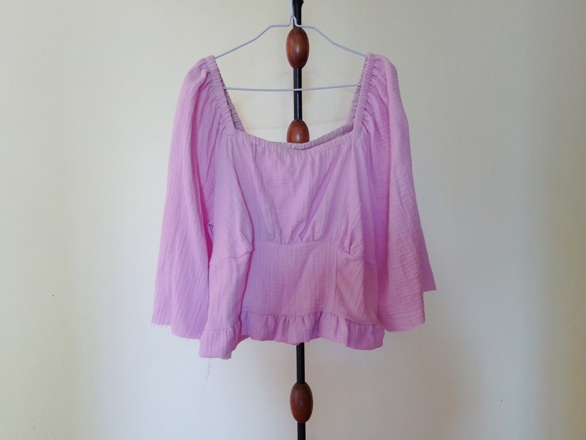 A lilac long-sleeved gathered top (Briarwood Top mock up) with a square neckline and ruffled hem, hanging on a hanger on a stand.