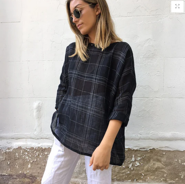 A woman wearing sunglasses and a loose-fitting, dark plaid blouse with drop shoulders and three-quarter sleeves, paired with white pants.