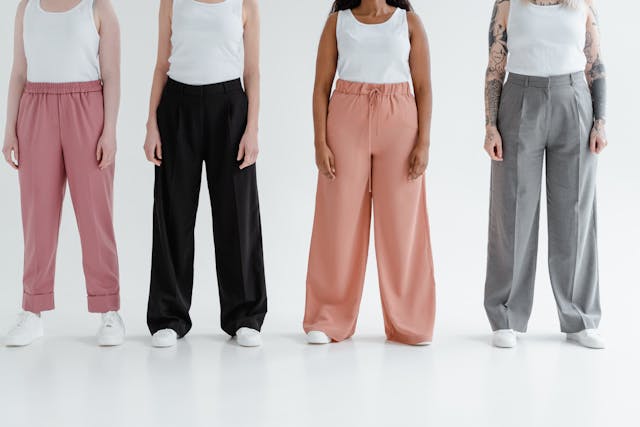 Four people standing in a line, each wearing a white tank top and different styles and colors of wide-legged trousers (pink elasticated, black pleated, peach drawstring, and grey pleated).