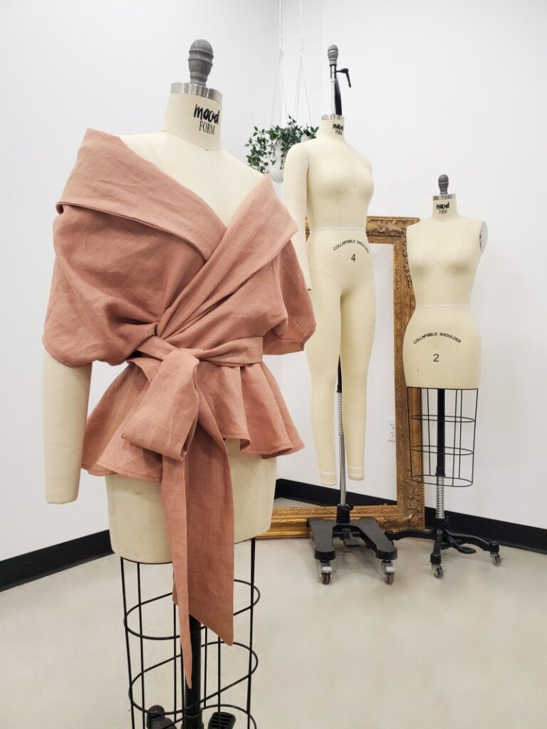 A dress form modeling an oversized, off-the-shoulder blouse made of rosy-pink linen, featuring a dramatic wrap design that ties into a large bow and flares into a peplum at the waist.