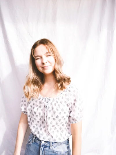 A smiling woman wearing a white, short-sleeved peasant blouse with a small black print and a gathered neckline with a center tie, tucked into blue jeans.